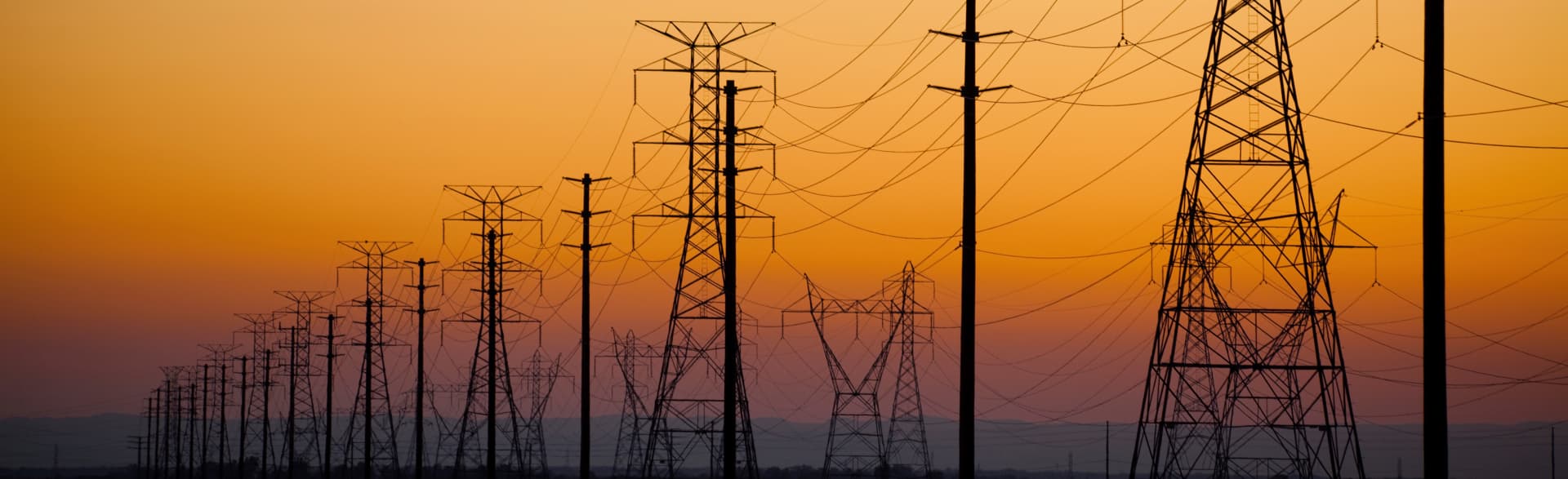 High-voltage transmission towers and power lines silhouetted against an orange dusk sky, from the Nexans website