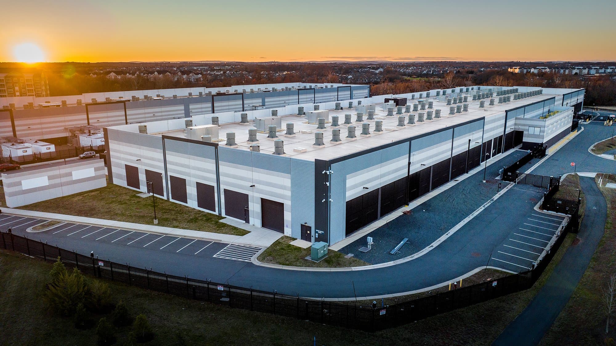 Aerial view at sunset of a large Amazon Web Services data center building with rooftop cooling fans and a gated access road