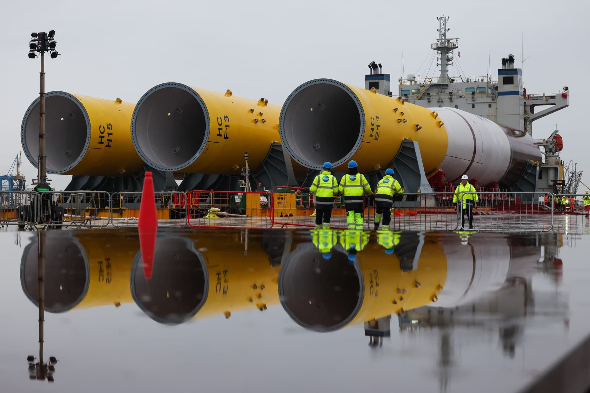 Yellow monopile foundations for the Hornsea 3 offshore wind farm on a ship at a UK port, with workers in hi-vis