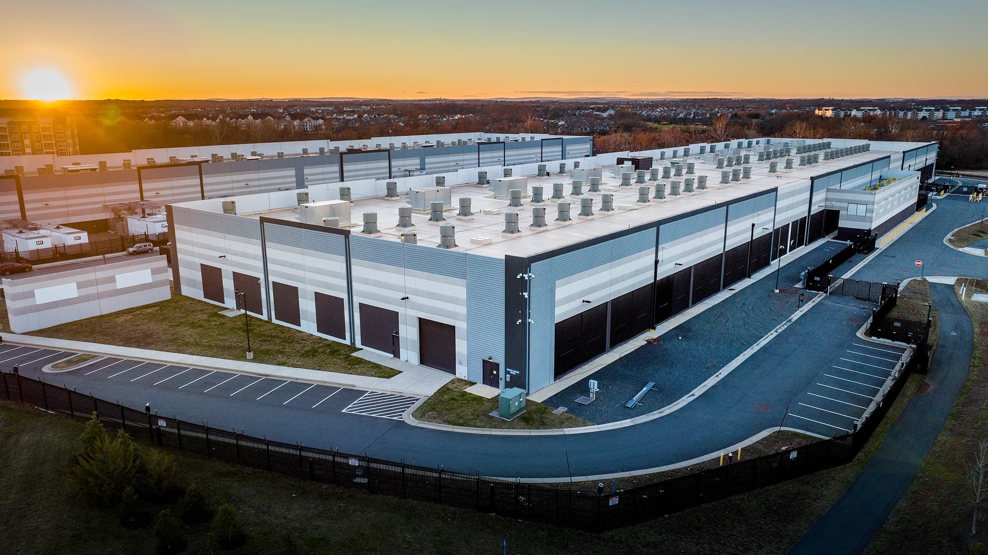Aerial view of an Amazon Web Services data center building at sunset, with rooftop cooling fans and a perimeter fence